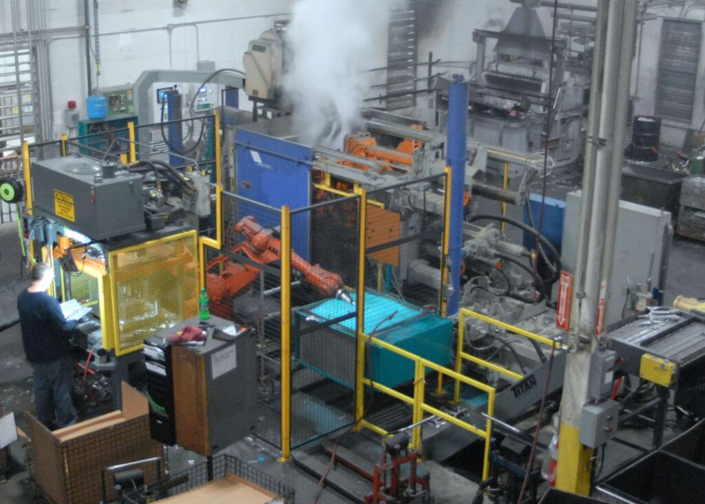 Worker operating machinery inside a large die casting manufacturing facility with automated equipment and steam.